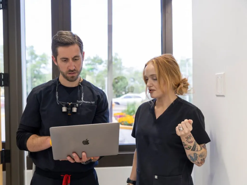 A dentist holding a laptop with a dental assistant in the hallway